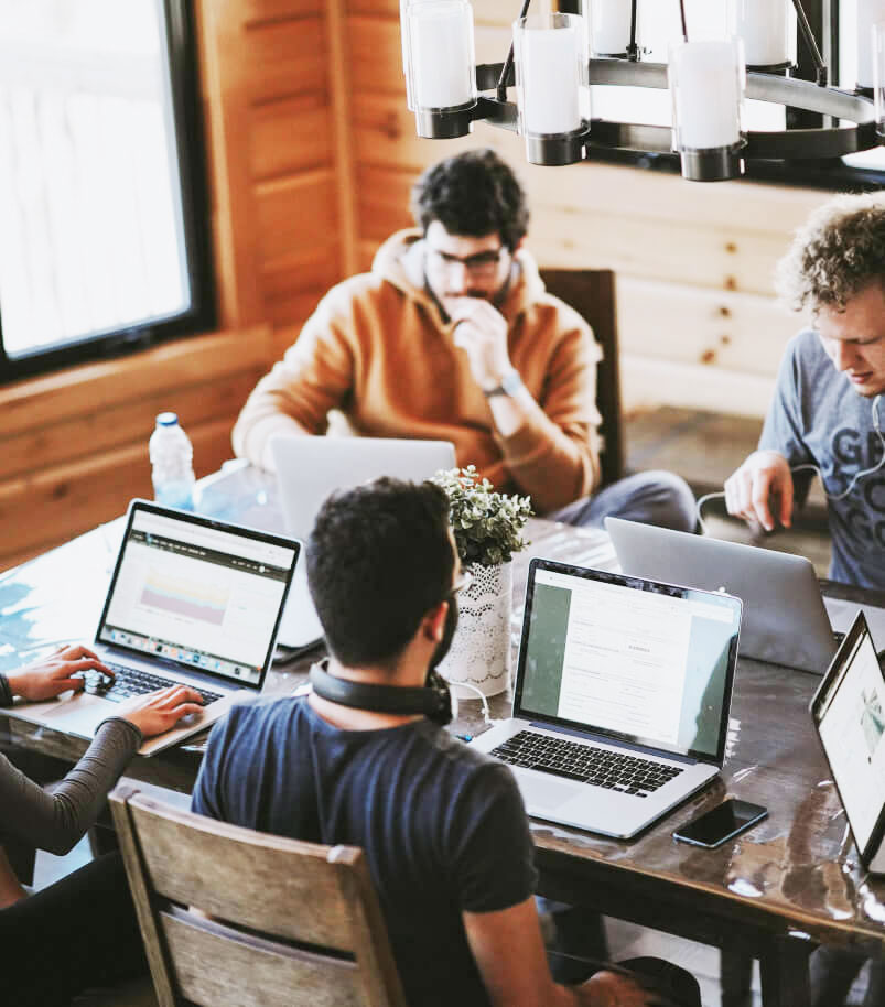 Group of people working together on laptops around a rustic wooden table in a modern, cabin-style room